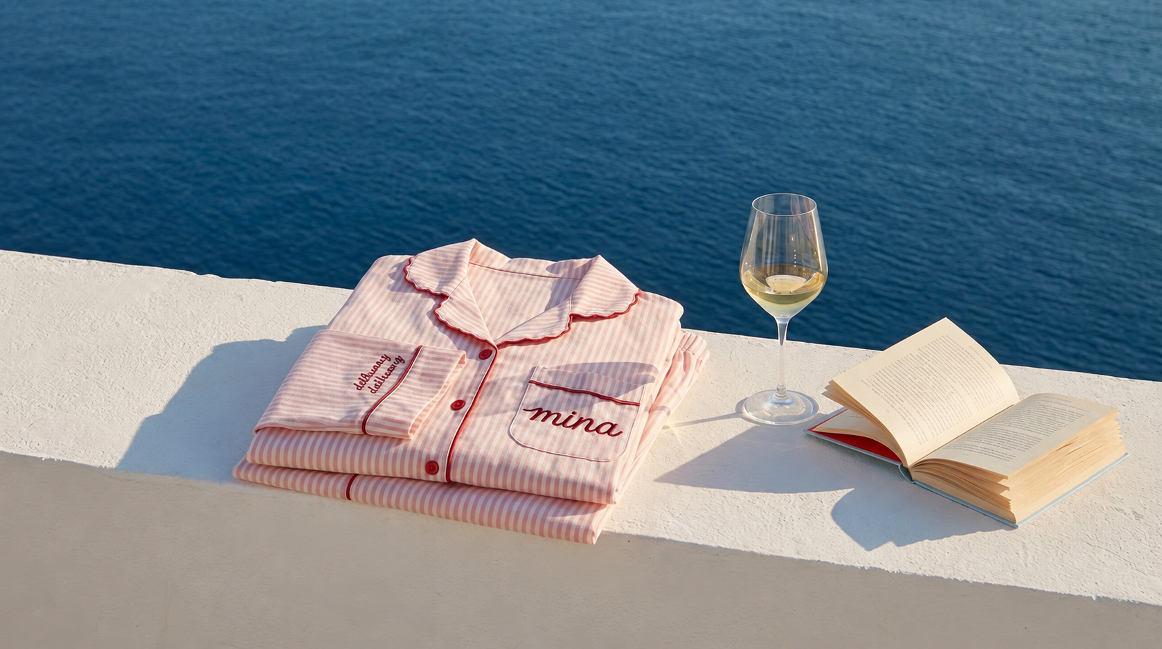 Personalized pink striped pajamas beside a book and wine glass on a Mediterranean terrace overlooking the sea